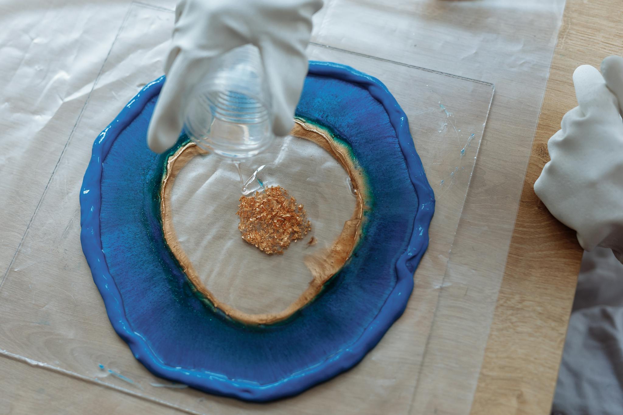 Close-up of resin art creation with blue and glitter accents being poured by gloved hands.